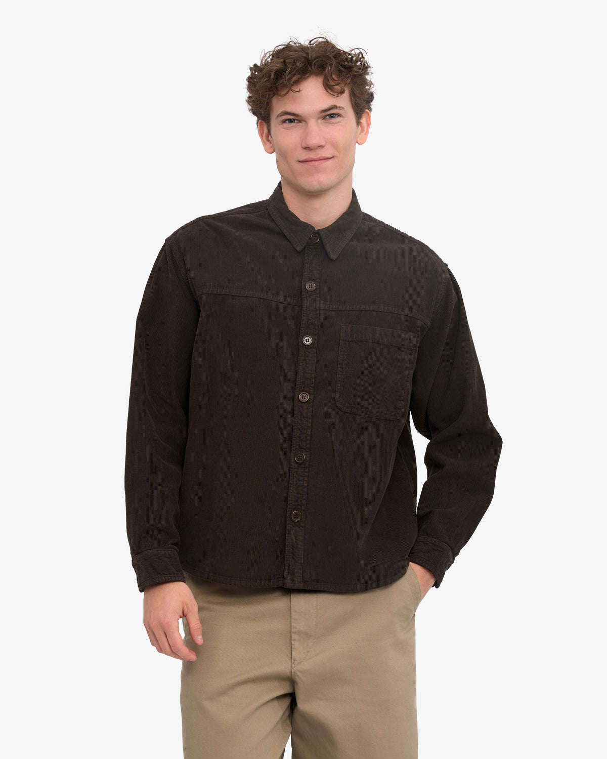 A man wearing a dark brown corduroy shirt with a single chest pocket, paired with khaki pants, standing against a light grey background.