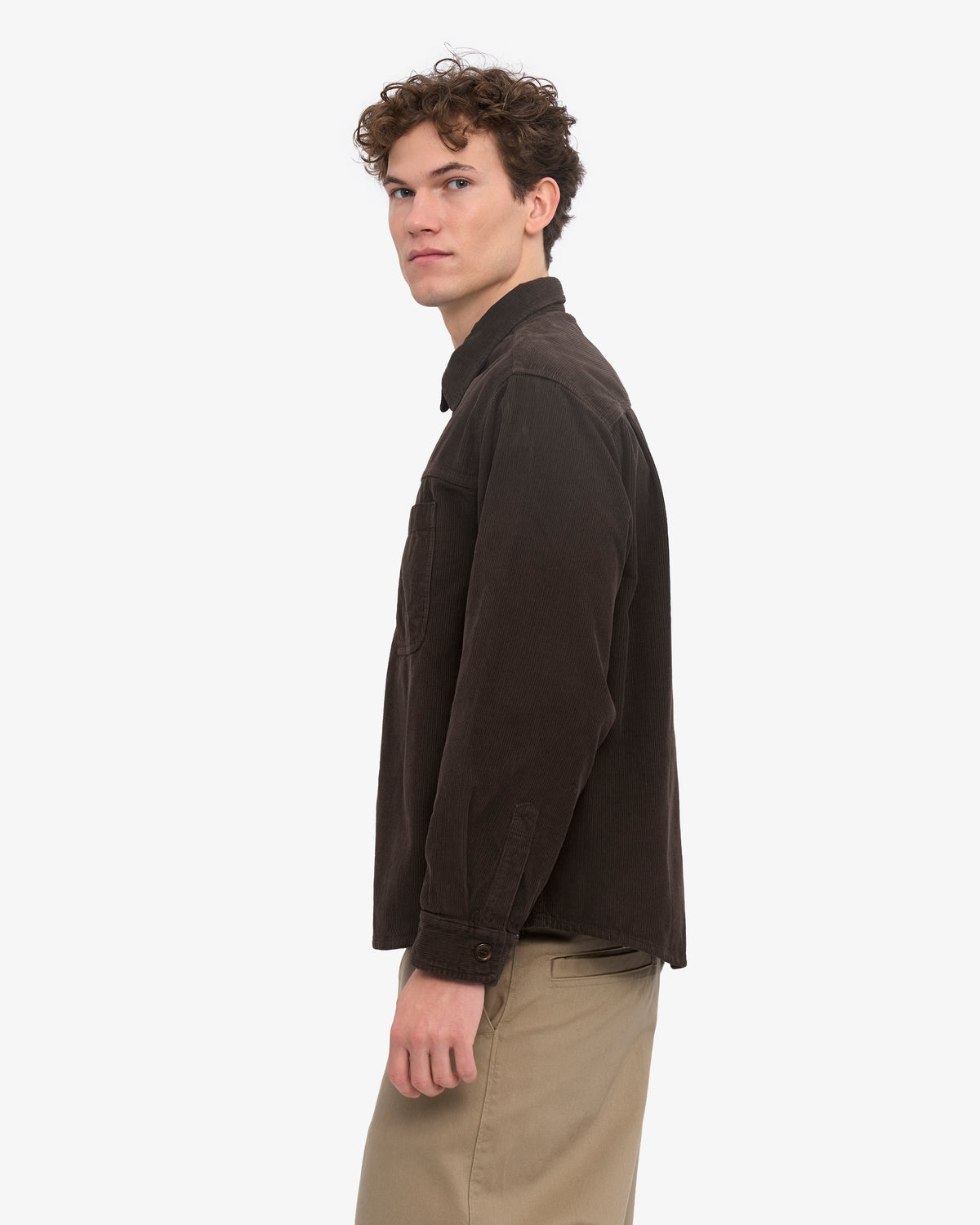 A man wearing a dark brown corduroy button-up shirt and beige trousers, shown in a side profile against a light gray studio background.